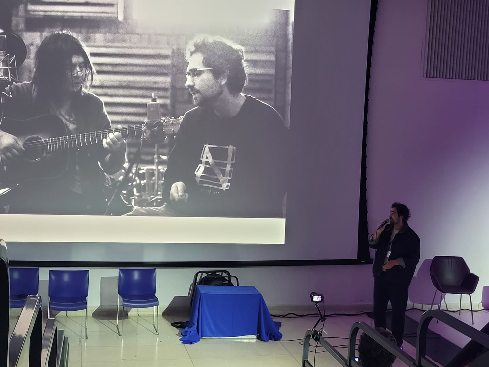 Adam Fainman speaking on the VRTO 2026 mainstage with a black and white photo of himself performing on screen behind him